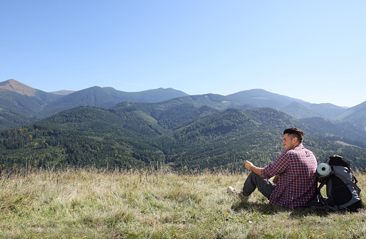 Young man enjoying the wonderful spring sunny day having picnic on the fresh green flowering meadow under the panoramic picturesque blue cloud sky in the mountains