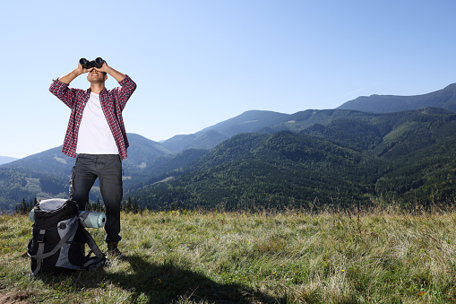 Tourist with hiking equipment looking through binoculars in mountains