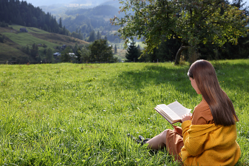 Young woman reading book on green meadow in mountains, back view