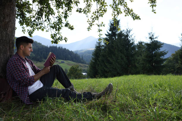 istock Handsome man reading book under tree on meadow in mountains 1400289632