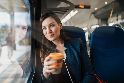 A beautiful young business woman is sitting in the cab of a high-speed train, looking through window and drinking coffee to go.