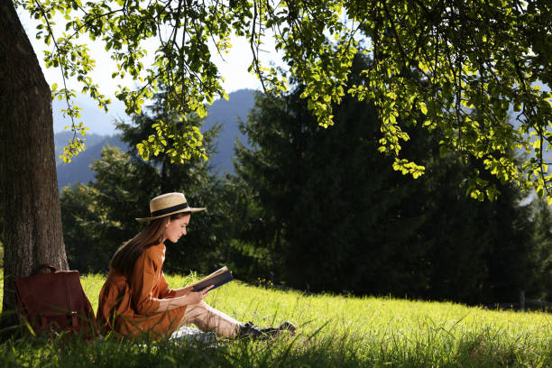 istock Young woman reading book under tree on meadow in mountains 1400290546