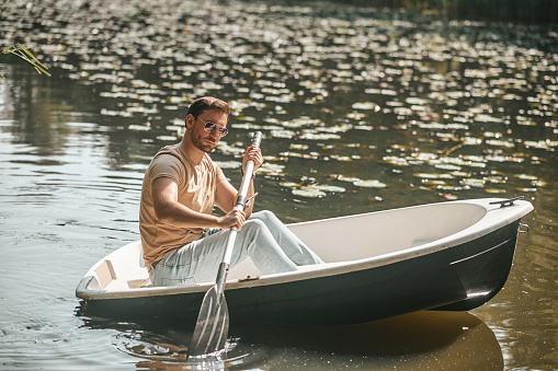 Serious focused dark-haired male in sunglasses seated alone in the boat rowing with one oar