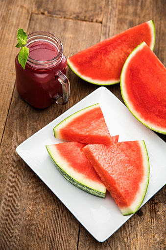 This is an outdoor photograph of a plate of sliced triangle watermelon sitting on a plate with a glass of smoothie watermelon juice and mint
