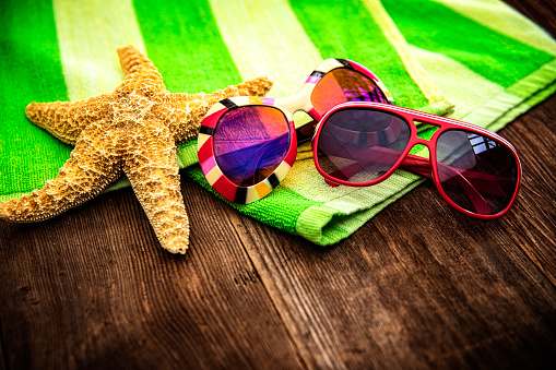 This is a close up photograph of a pair of sunglasses. One is for men the other is for women sitting on top of a green striped beach towel on a wood deck. There is space for a copy