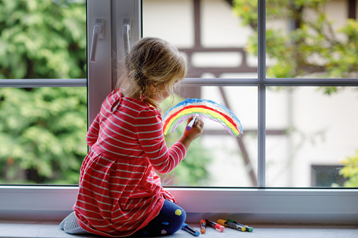 Adoralbe little toddler girl with rainbow painted with colorful window color during pandemic coronavirus quarantine. Child painting rainbows and hearts around the world with words Let's all be well