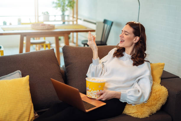 Attractive woman having fun at home. Eating pop corn and watching movie on laptop. Young woman sings for fun.jpg stock photo