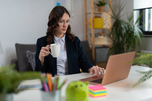 Beautiful businesswoman holding cup of coffee and looking at laptop in office stock photo