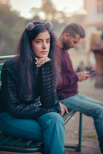 In this outdoor day image with copy space, an Asian/Indian sad young woman contemplates sitting on a park bench with a man who uses a smartphone.