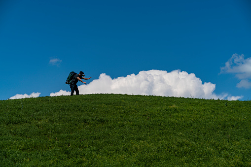 mercimek tarlası olan yeşil tepe, tepenin arkasından yükselen beyaz bulutlar ve mavi gökyüzü arka planı. tepenin zirvesinde bulutlar önünde sırt çantalı gezgin adam yürüyor. çevre temalı bu fotoğraf full frame makine ile çekilmiştir.