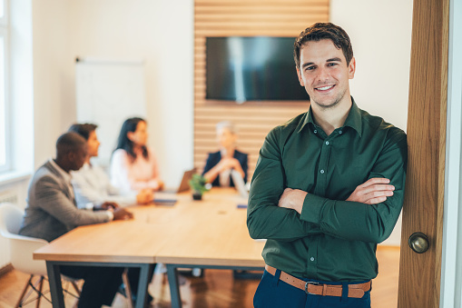 Shot of a businessman standing in the office with his arms crossed looking confident and smiling at the camera, colleagues behind