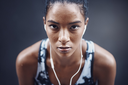 Portrait of one active young mixed race woman with sweat on her face wearing earphones and taking a rest break after run or jog exercise outdoors. Focused female athlete looking tired but determined after challenging workout against a dark background