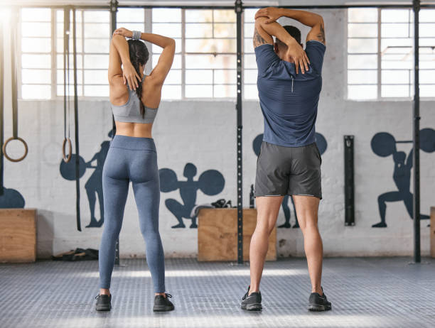 due atleti caucasici attivi da dietro allungano le braccia e i tricipiti tirando il gomito con le mani verso la colonna vertebrale dietro la testa mentre si esercitano in palestra. uomo e donna che preparano il corpo per l'allenamento di allenamento in un  - stretching back rear view women foto e immagini stock