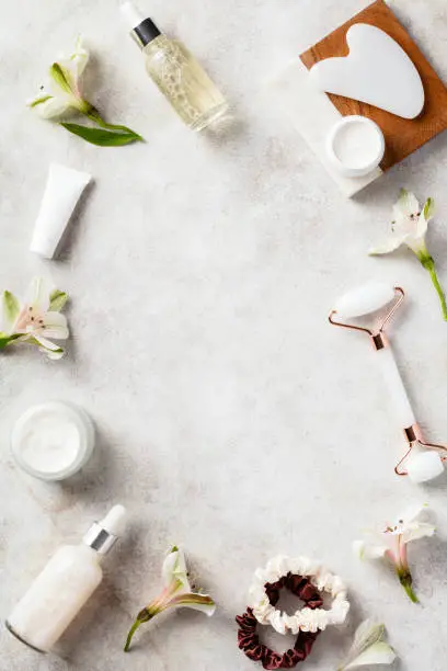 Photo of Flat lay composition made of natural skincare cosmetics, gua sha, face massage roller, flowers on stone table.