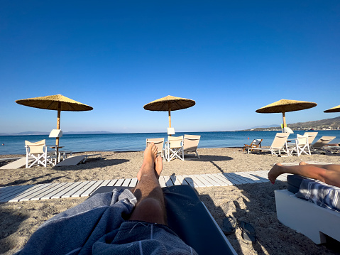 Summer vacation travel concept: Male legs relaxing on a sun bed under the shadow of parasol. Beautiful empty beach with lined sun chairs and umbrellas on sunny day. Waiting for hotel guests, travelers. Holiday background with large copy space.