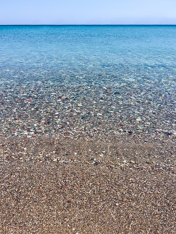 Summer vacation travel concept: Beautiful tranquil crystal clear water surface. Horizon over water. Pebble beach. Blue ocean and sky background with large copy space. Ripples, sun glare, movement on water