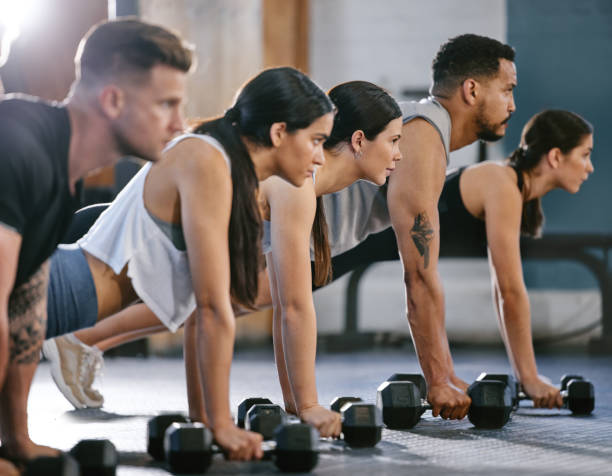 istock Diverse group of active young people doing plank hold and push up exercises with dumbbells while training together in a gym. Focused athletes doing press ups and renegade rows with weights to build muscle and endurance during a workout in a fitness class 1400315443