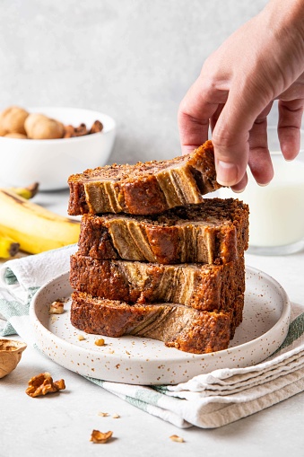 Woman taking slice of tasty banana bread with chopped walnuts, chocolate and cinnamon and milk on light background. Stack of pieces of bread. Breakfast concept. Selective focus.