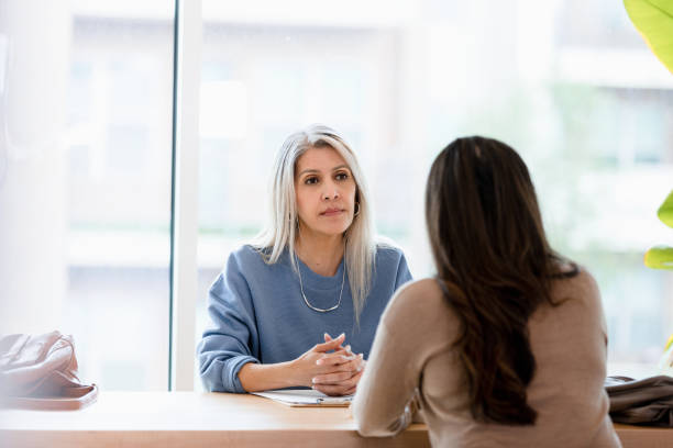 Serious businesswoman listens to unrecognizable female client With her hands clasped in front of her, the mature adult businesswoman listens with a serious look on her face to the unrecognizable female client. grave stock pictures, royalty-free photos & images
