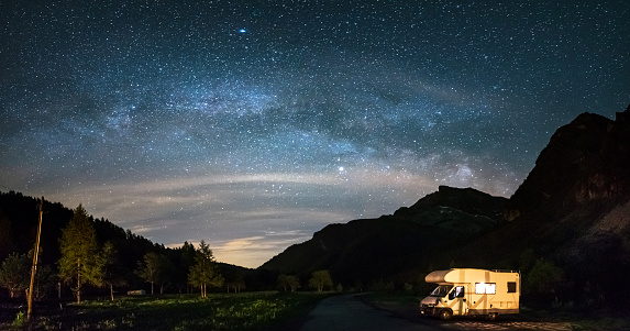 Camper van under panoramic night sky in the Alps. The Milky Way galaxy arc and stars over illuminated motorhome. Camping freedom in unique landscape.