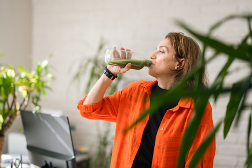 A businesswoman is having smoothie in her environmentalist green office with lots of house plants and sunlight from windows.