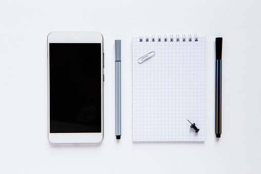 Minimalistic monochrome flat lay of a working space with white blank notebook page, mock up phone, pens, button and clip on white background. Business office or planning concept. Top view.