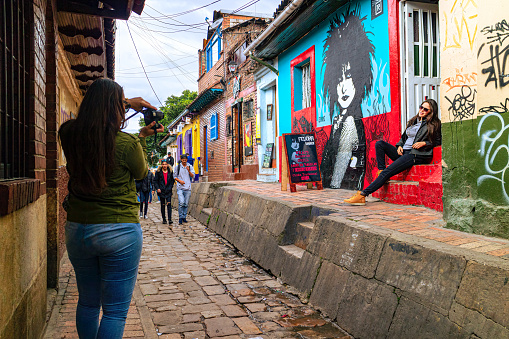 Bogota, Colombia - October 20, 2019: Both Tourists and local Colombian people walk up the narrow Calle del Embudo one of the most colorful streets in the historic La Candelaria district of Bogotá, the Andean capital city of the South American country of Colombia. The street leads to the Chorro de Quevedo, the plaza where it is believed the Spanish Conquistador, Gonzalo Jiménez de Quesada founded the city in 1538. Many street facing walls in this area are painted with either street art or the legends of the pre-Colombian era, in the vibrant colours of Colombia. The altitude at street level is 8,660 feet above mean sea level. Photo shot on an overcast morning; horizontal format.
