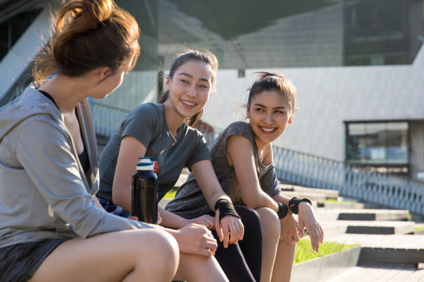 Three young ladies sitting on the steps of a modern building in the city on a sunny summer day - stock photo Suitable for sportswear, sports equipment, sports drinks, sun protection products, youth culture, smartphone applications, sports products, urban sports, girls' products, fitness places, sports and leisure, print advertising. Portrait. central asian ethnicity photos stock pictures, royalty-free photos & images