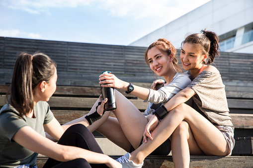 Three young ladies sitting on the steps of a modern building in the city on a sunny summer day, they pass sports water bottles - stock photo