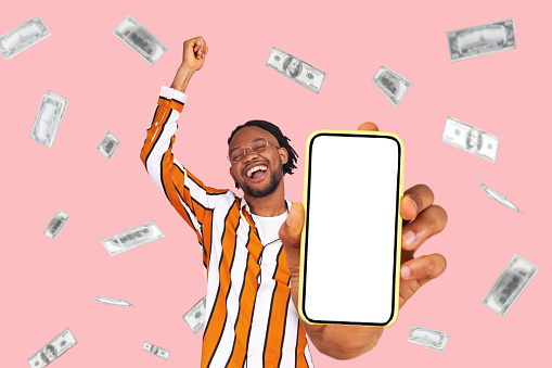 Online investment, winner overjoyed rich man and dollars money earning. Portrait of amazed young man showing mobile empty display and celebrating his victory. indoor isolated on pink background.