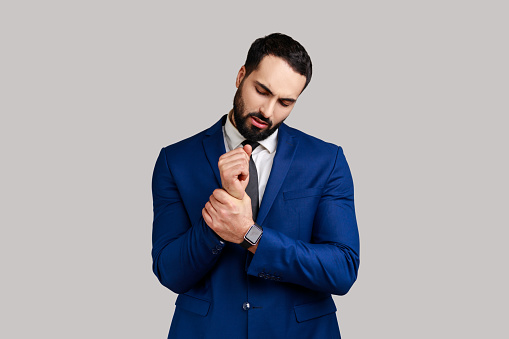 Portrait of businessman standing with grimace of pain, massaging sore wrist, suffering hand injury or sprain, wearing official style suit. Indoor studio shot isolated on gray background.
