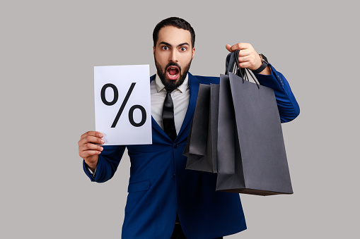 Shocked astonished bearded businessman holding shopping bags in hands and paper with percent mark, big discounts, wearing official style suit. Indoor studio shot isolated on gray background.