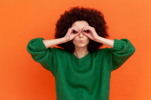 Funny woman with Afro hairstyle wearing sweater looking through fingers in binoculars gesture, observing distant with attentive look, watching afar. Indoor studio shot isolated on orange background.