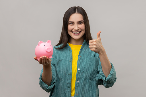 Portrait of smiling pleased woman with dark hair holding pig money box in hands, saving, showing thumb up, wearing casual style jacket. Indoor studio shot isolated on gray background.