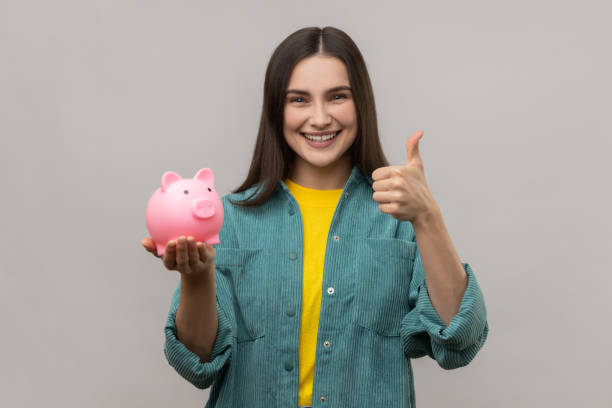 Pleased woman with dark hair holding pig money box in hands, saving, showing thumb up. stock photo