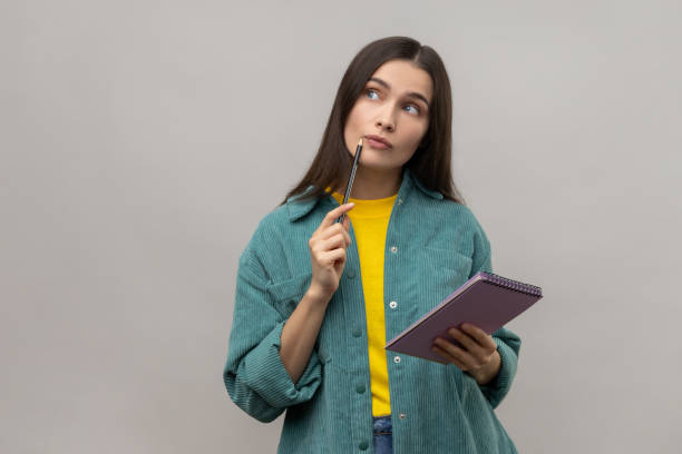 istock Thoughtful woman standing with paper notebook and pen, looking away with pensive expression. 1400354508