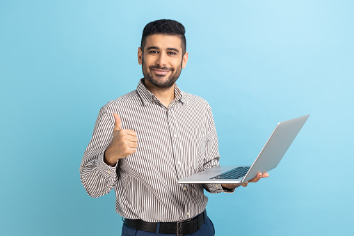 Portrait of smiling positive businessman standing with portable computer in hand, looking at display, showing thumb up, wearing striped shirt. Indoor studio shot isolated on blue background.