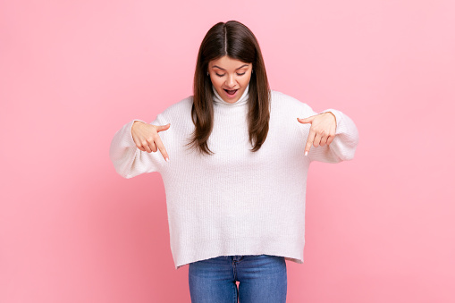 Excited, surprised girl pointing down and looking with shocked face, showing place for commercial, wearing white casual style sweater. Indoor studio shot isolated on pink background.