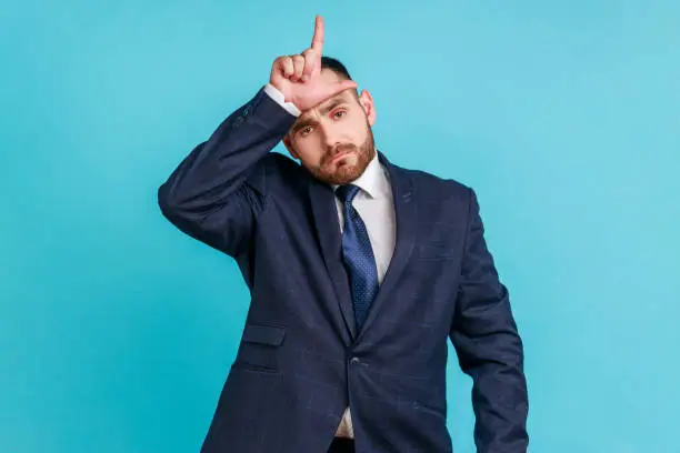 Portrait of upset disappointed young adult businessman with beard in dark suit standing, showing looser gesture with hand on forehead. Indoor studio shot isolated on blue background.