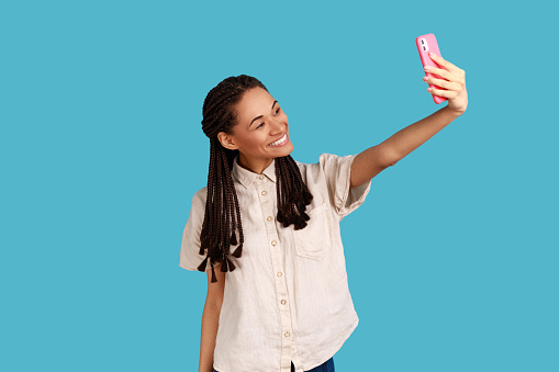Young lovely woman with black dreadlocks looks at camera of smartphone, takes selfie portrait, broadcasting livestream, wearing white shirt. Indoor studio shot isolated on blue background.
