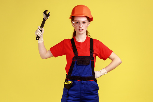 Portrait of confident builder woman holding adjustable wrench, looking at camera with serious expression, wearing overalls and helmet. Indoor studio shot isolated on yellow background.