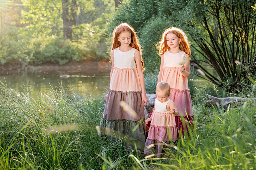 three red-haired sisters in long linen dresses are resting on the lake in the park on a sunny summer day.