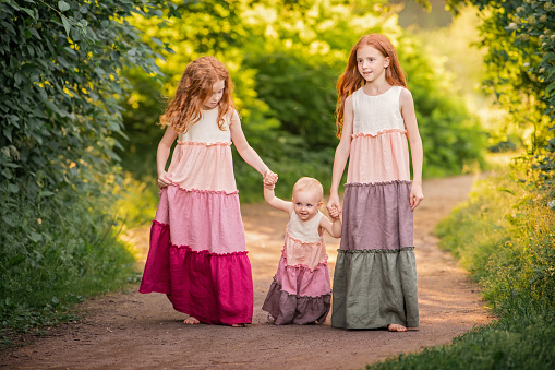 three red-haired girls in long dresses run barefoot along the path in the park towards the sunset, view from the back.