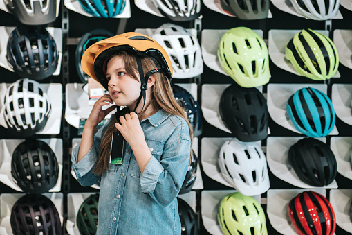 portrait of smiling happy little girl trying on cycling helmet in bike shop in front of shelf with lots of helmets