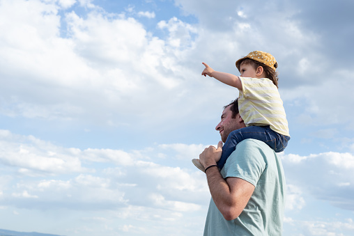 Side view of a smiling father carrying her little daughter on shoulders while she is pointing out.