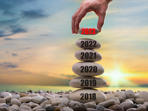 Happy new year 2023 concept. Entering the year 2023. Years ( 2018-2019-2020-2021-2022 ) written on the rising stone pile. Man hand adding stone to tower. Background is blurred sunset sky