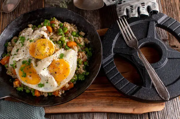 Healthy fitness or workout meal with fried eggs, brown rice and vegetables. Served in a rustic pan with dumbbell on wooden table background from above.
