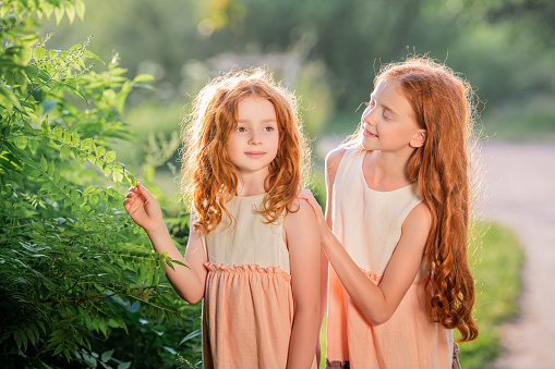 Two redhead sisters in long linen dresses are walking in the park on a sunny summer day.