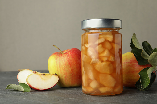 Tasty apple jam in glass jar and fresh fruits on grey table, space for text