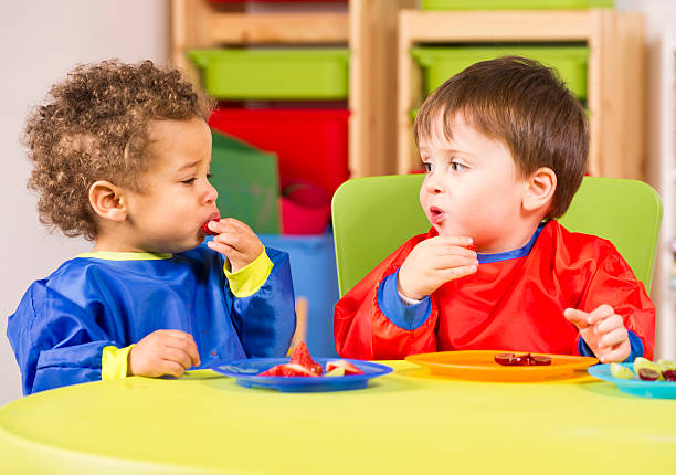two toddlers eating fruit in a nursery - jongen peuter eten stockfoto's en -beelden